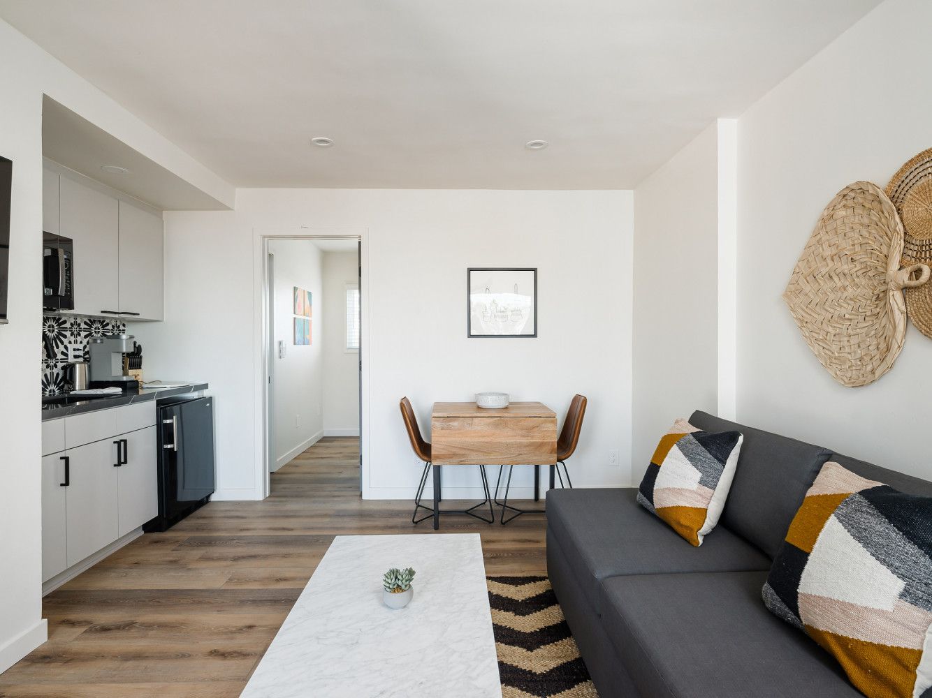 Bright modern living room with gray sofa, white rug, small dining table, and open kitchen.
