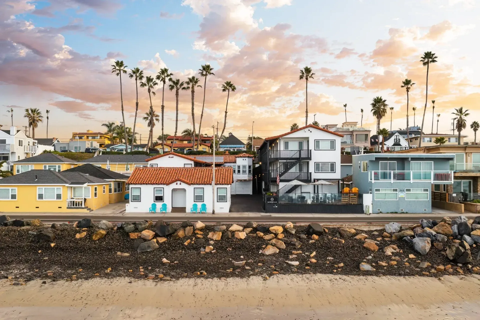 Beachfront homes behind a rocky seawall at sunset, with tall palm trees and pastel sky.