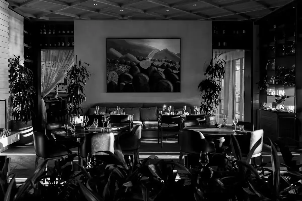 Dim restaurant dining room with tables, chairs, plants, and a large landscape painting on the wall.