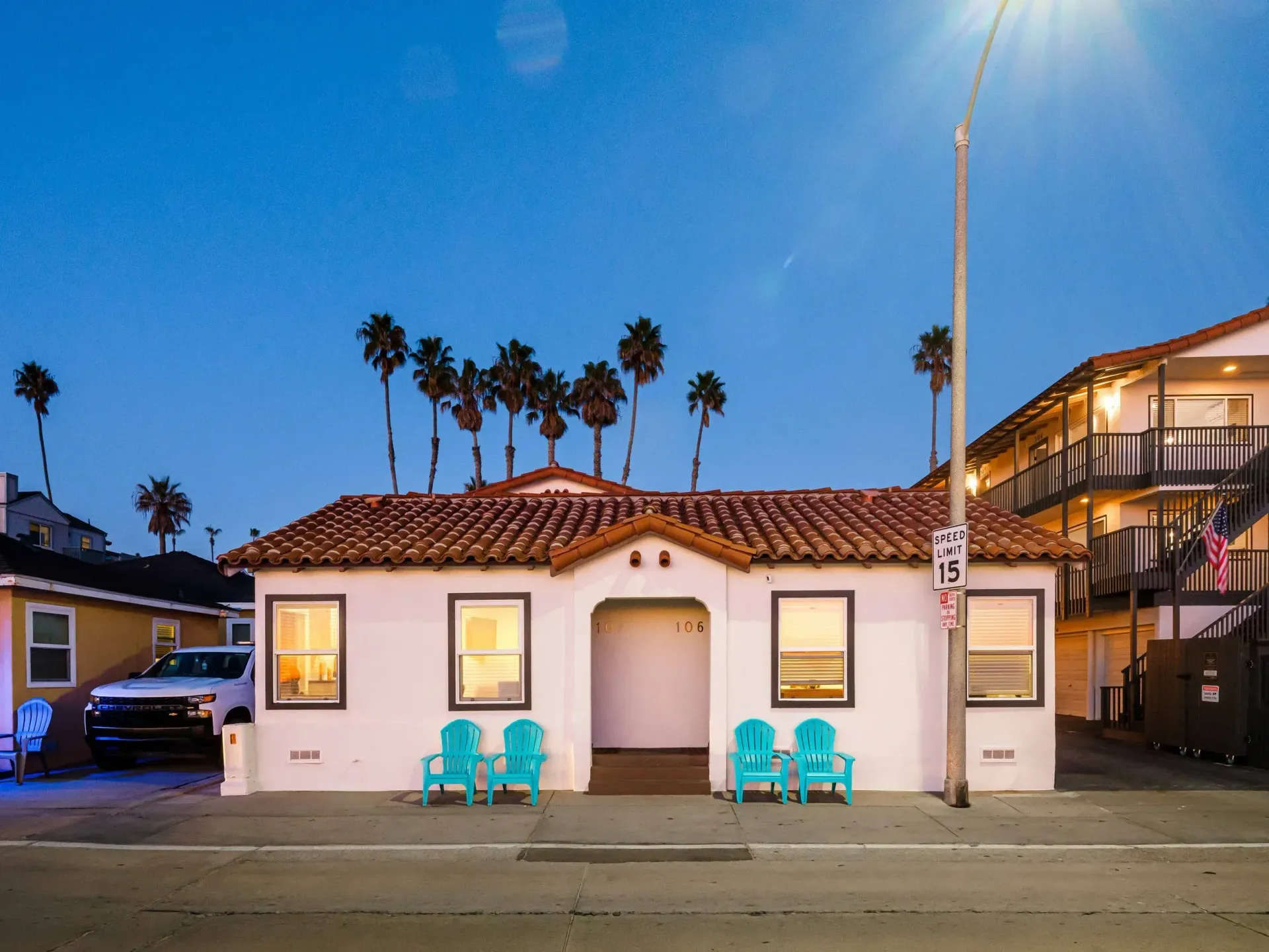 Small white stucco house at dusk with palm trees and turquoise chairs on the sidewalk