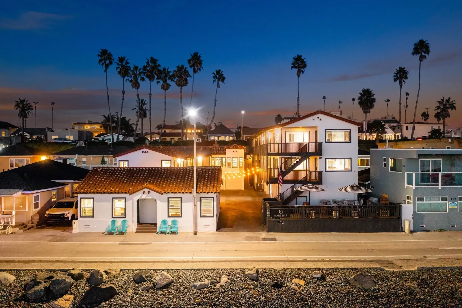 Row of beach houses at dusk with palm trees and warm lights along a coastal road