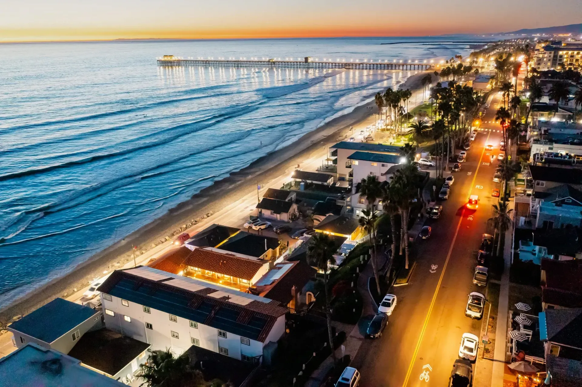 Aerial view of a coastal road at sunset beside the ocean, with a long pier and city lights glowing.