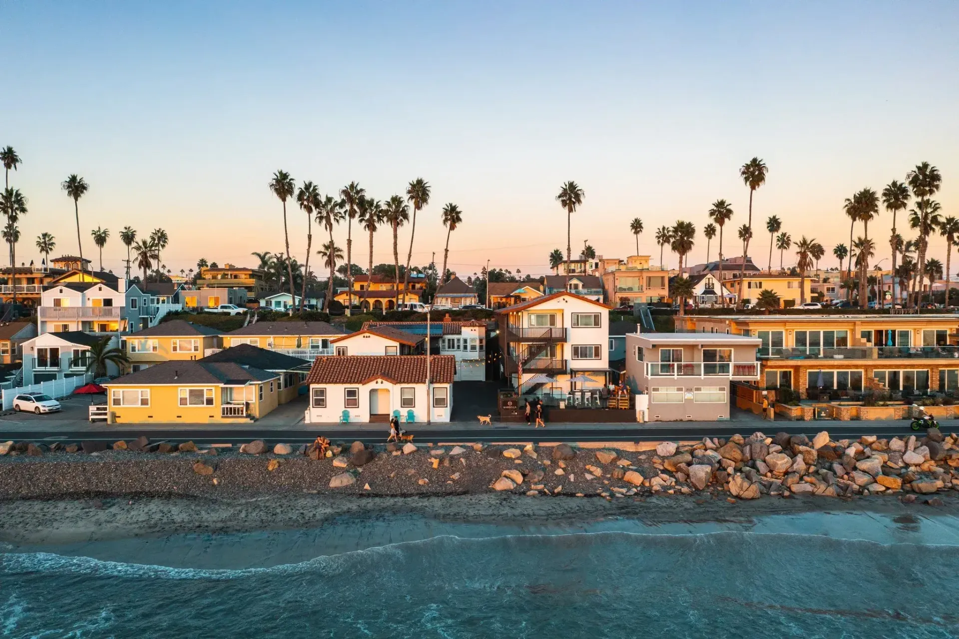 Coastal neighborhood at sunset with palm trees, houses, and rocky shoreline beside blue ocean waves
