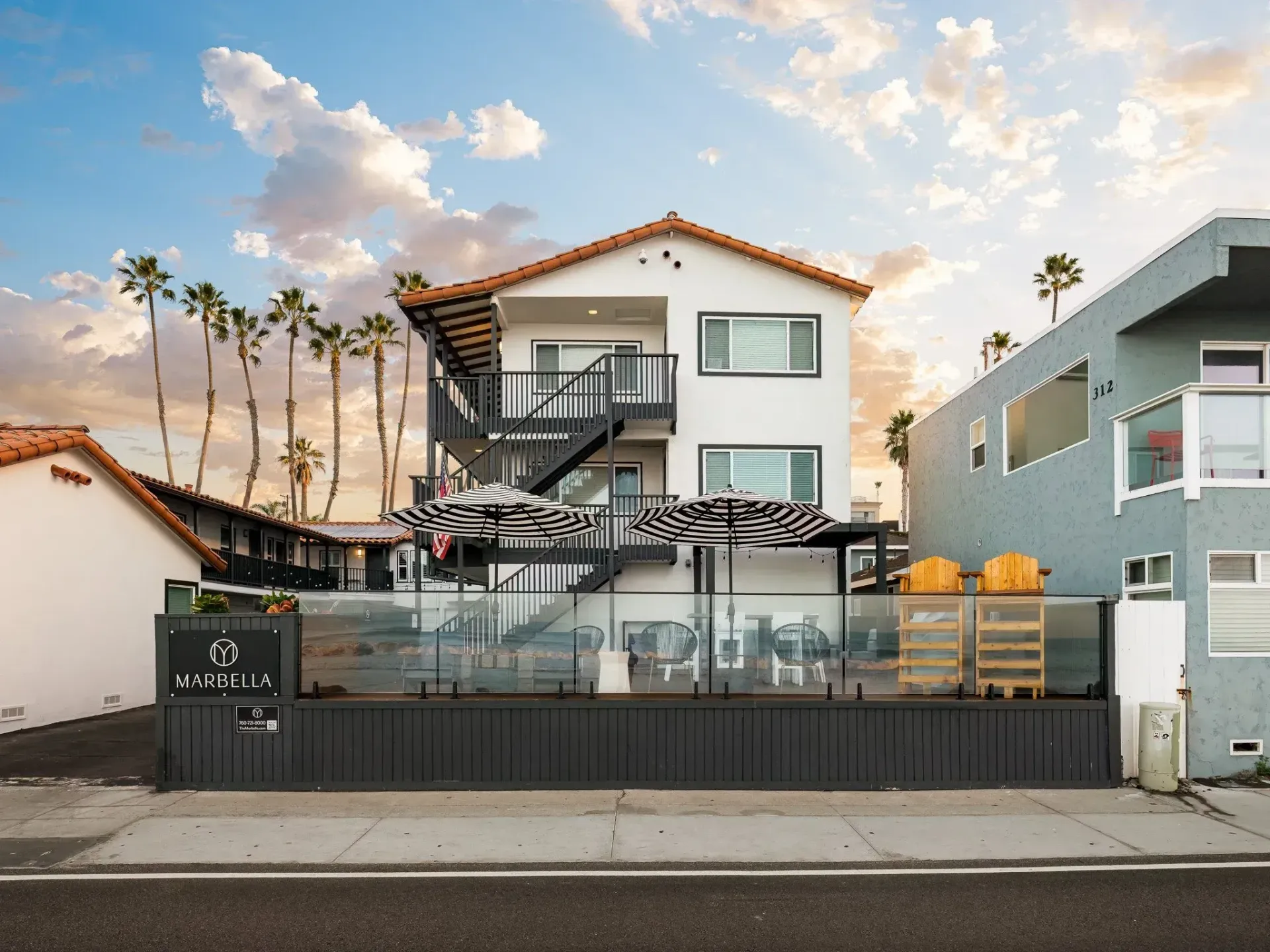 Modern white beach house with rooftop deck and pool, framed by palm trees at sunset