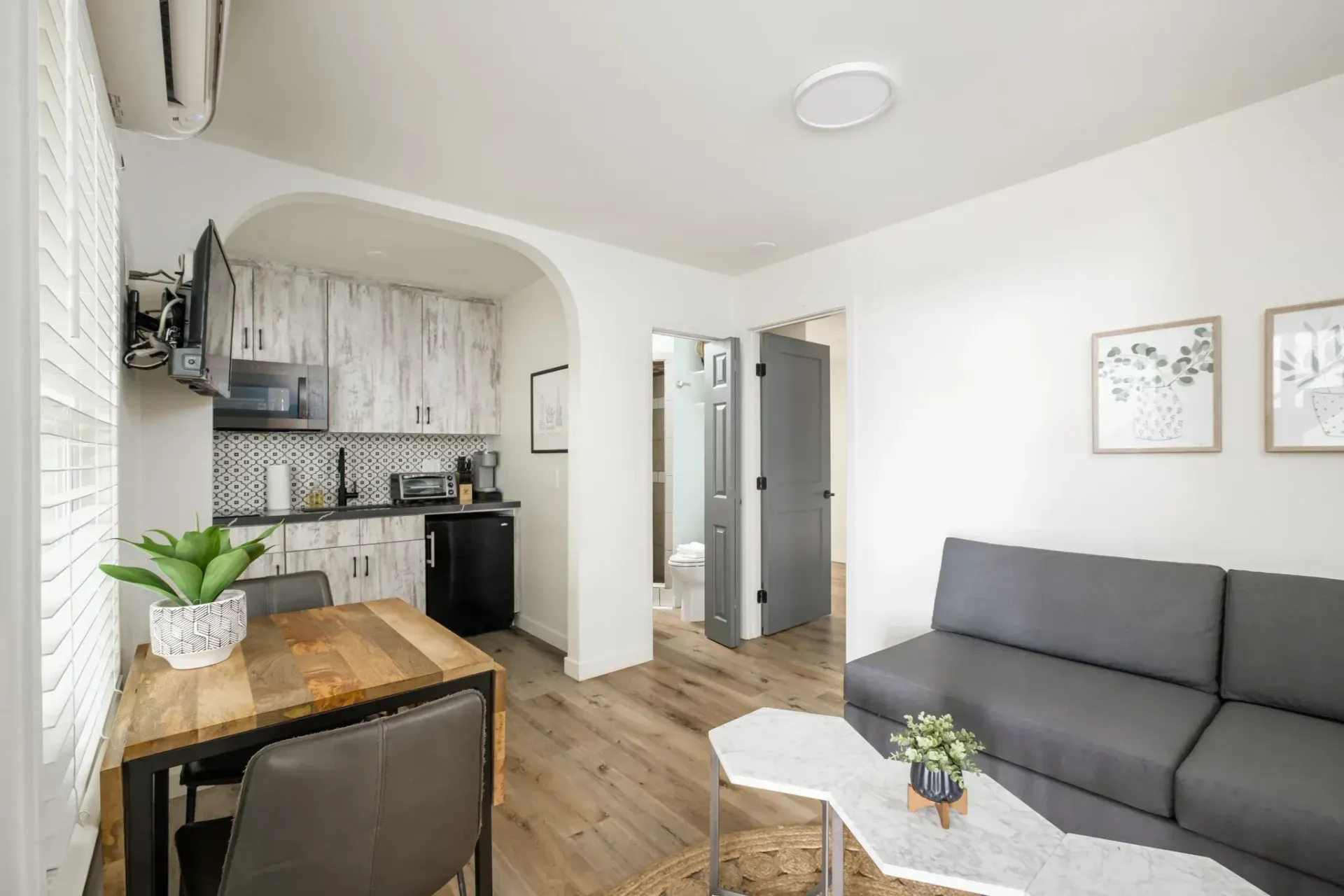 Modern living room with gray sofa, wood floor, small dining table, and compact kitchen area