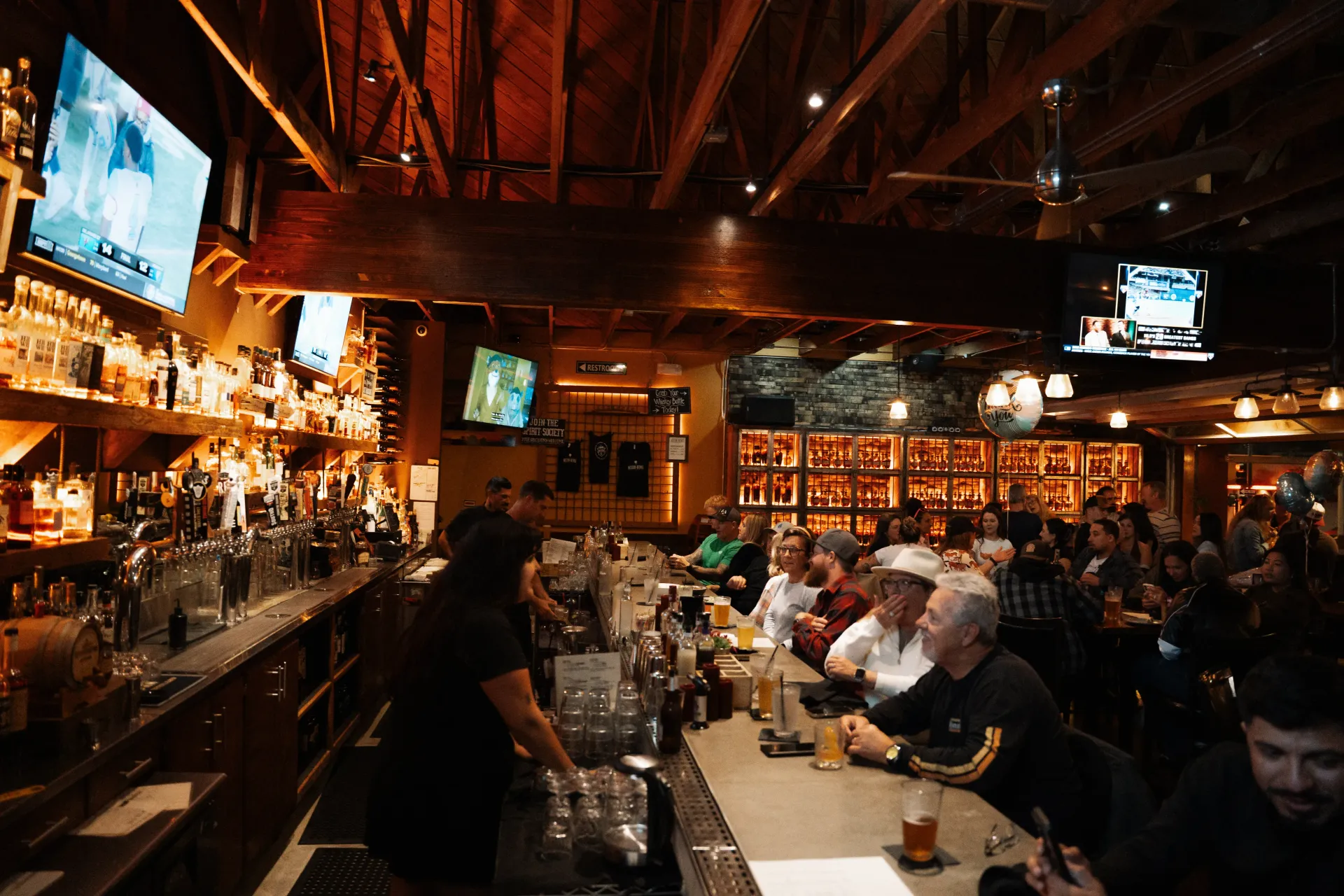 Crowded dimly lit bar interior with patrons seated at the counter and TVs above the shelves.