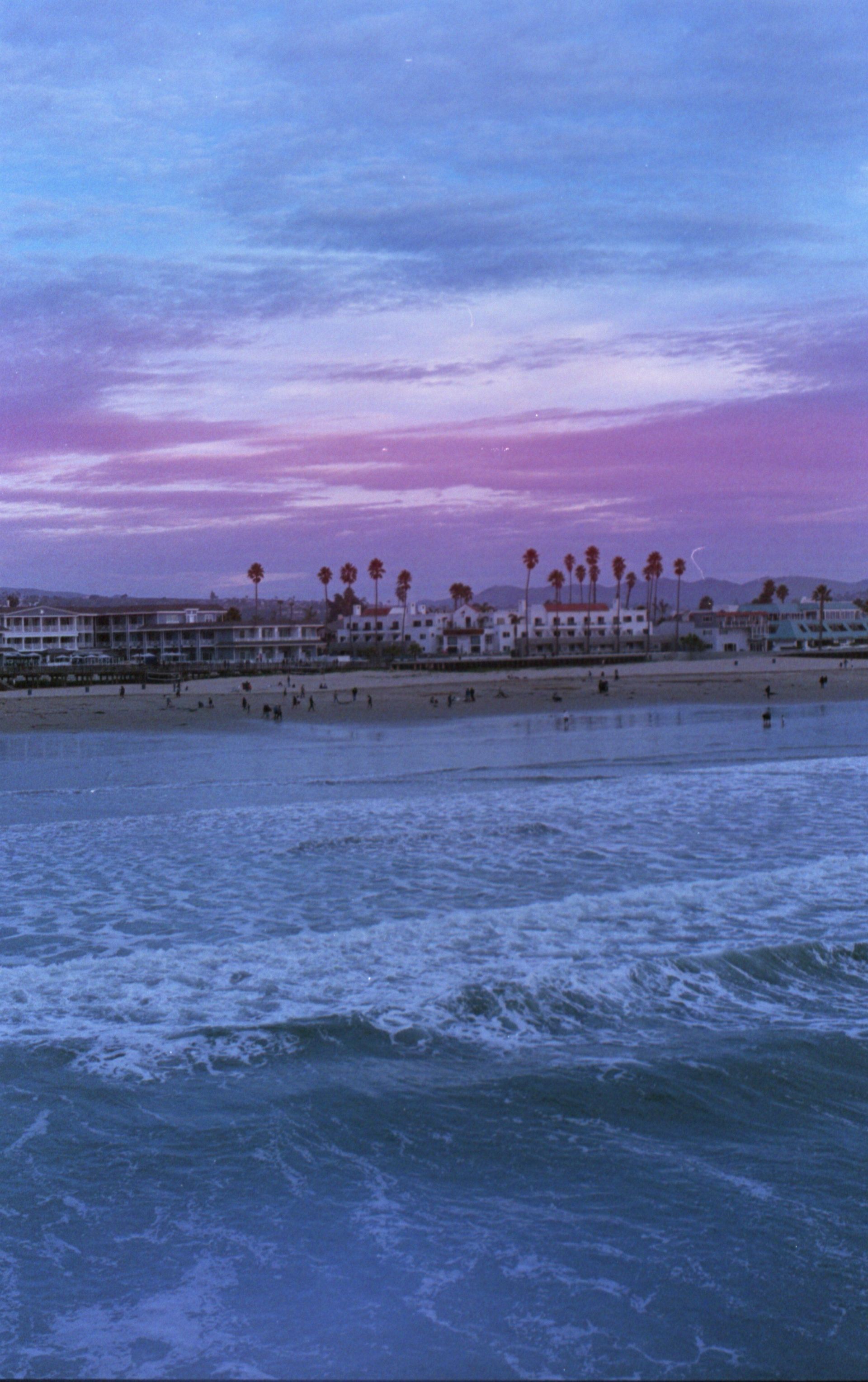 Purple sunset over a beach with gentle waves, palm trees, and buildings along the shore