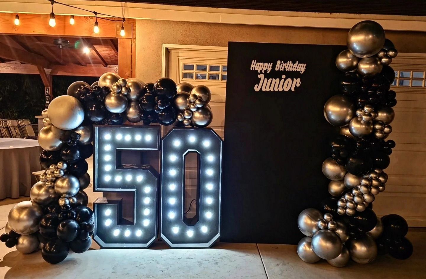 50th birthday setup with lit marquee numbers and black-silver balloon columns beside a “Happy Birthday Junior” sign
