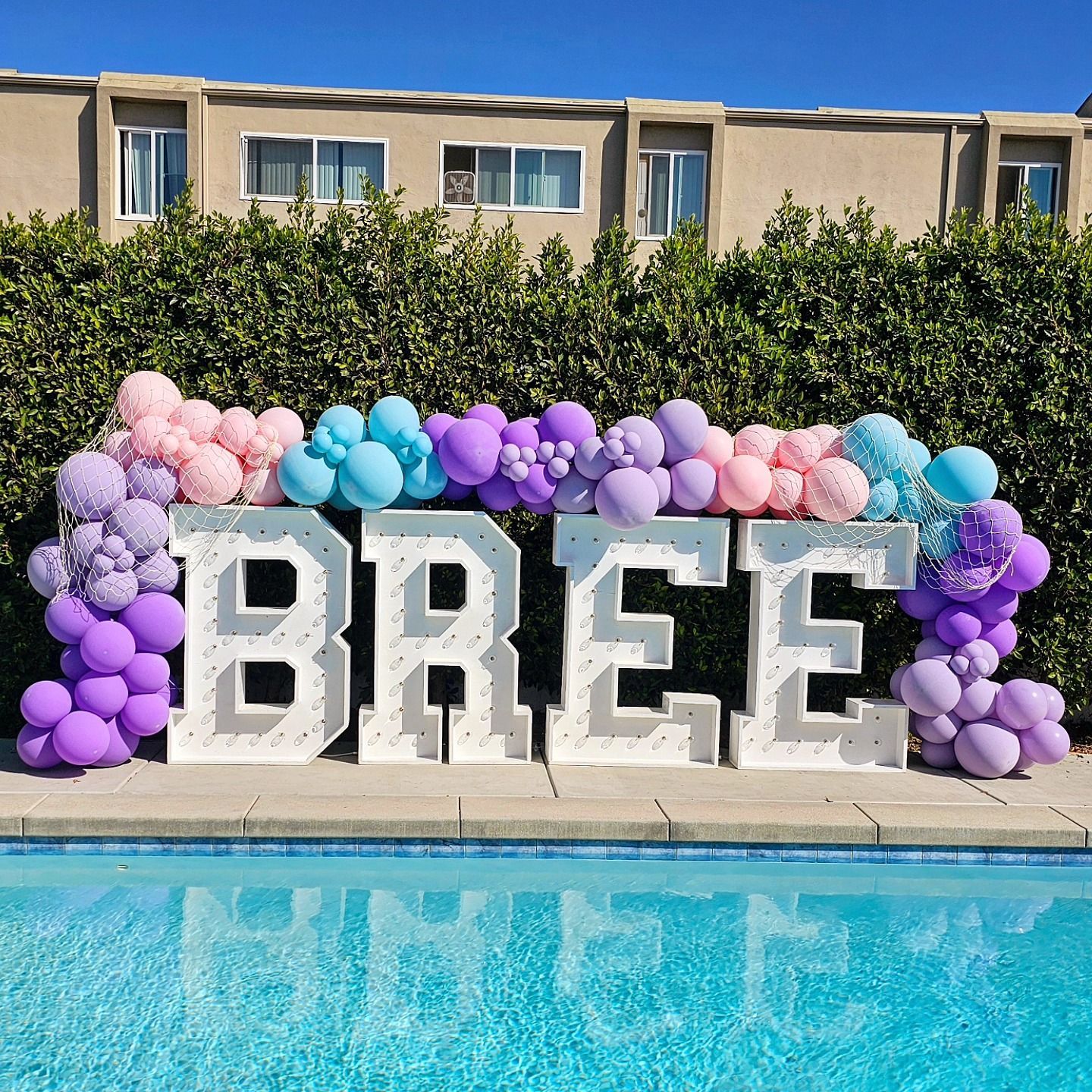 Colorful balloon arch with large white “BREE” letters beside a pool under a blue sky