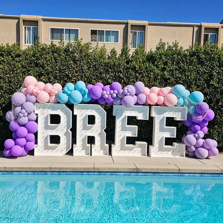 Large white “BREE” letters with pastel balloon arch by a pool under a clear sky