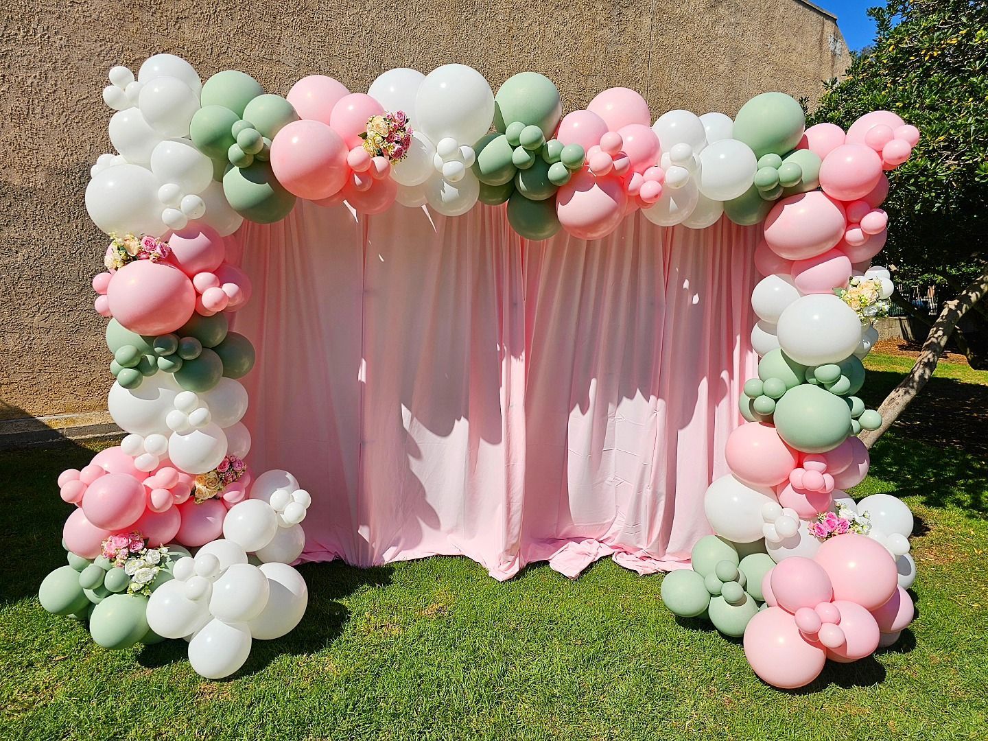 Pink and green balloon arch framing a pastel pink backdrop outdoors on grass