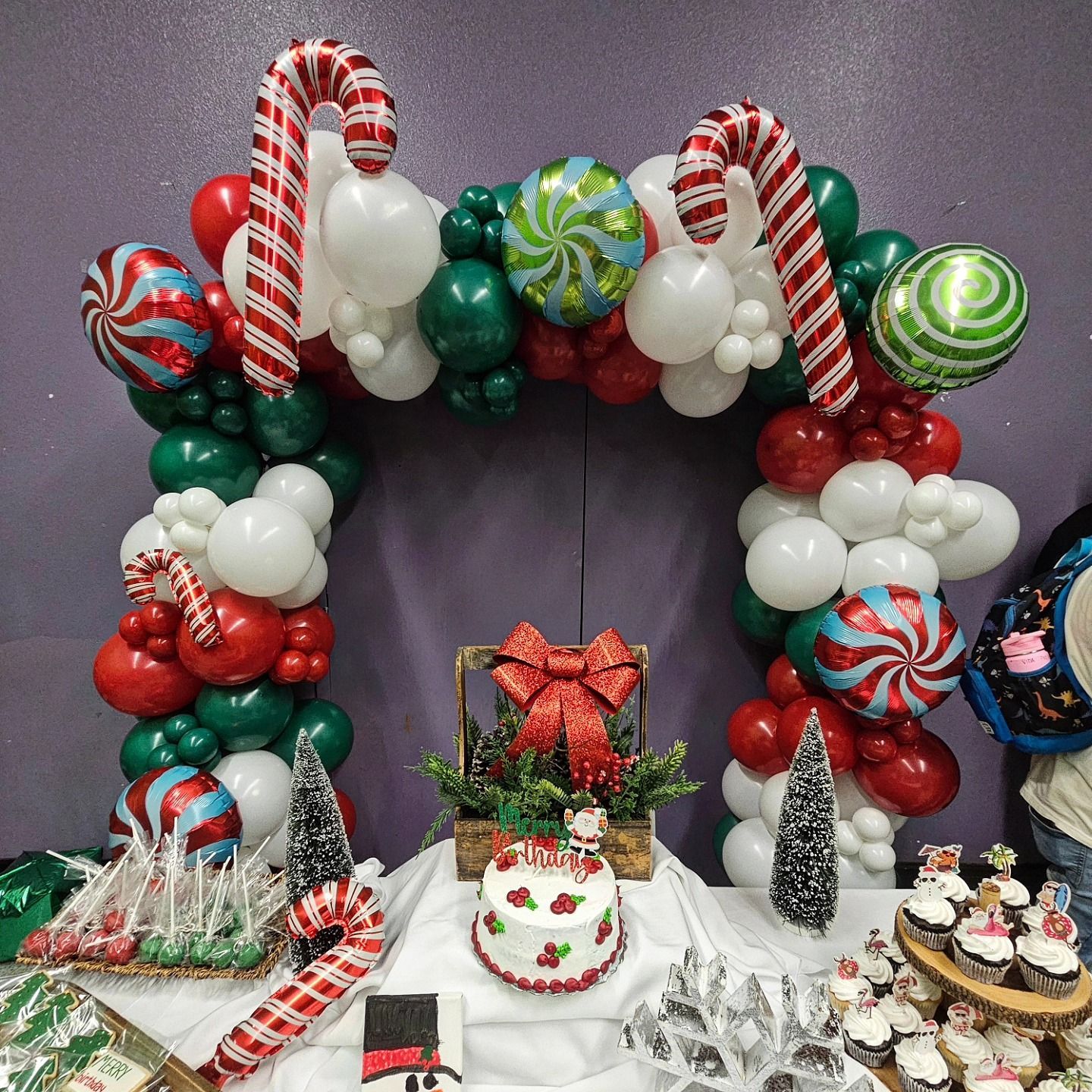Candy cane and ornament balloon arch over a holiday dessert table with cakes, treats, and Christmas décor