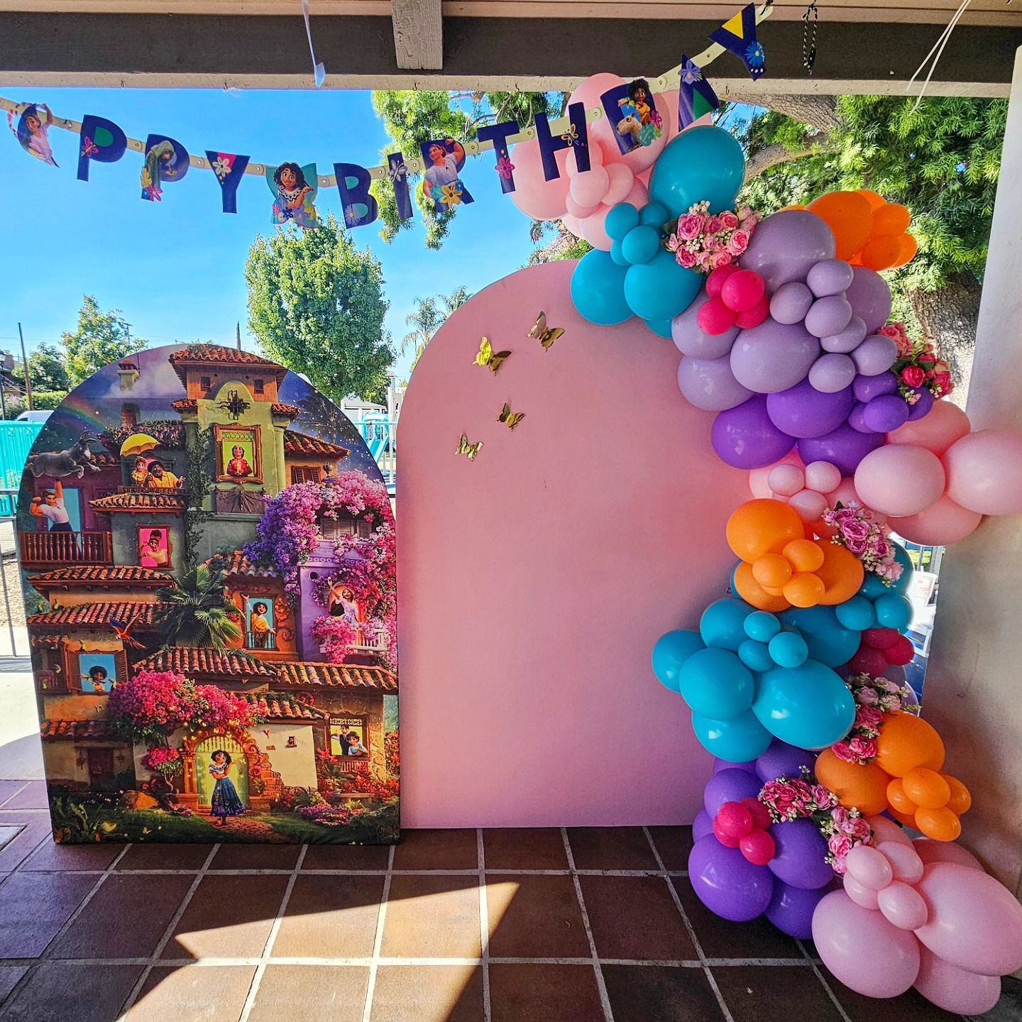 Colorful birthday party backdrop with balloons, “Happy Birthday” banner, and a pink display board outdoors