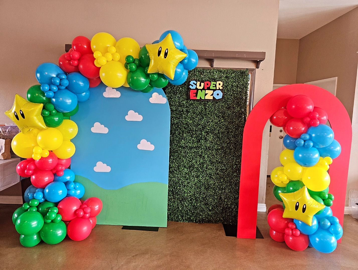 Colorful balloon arches frame a party backdrop with clouds, a green hedge panel, and a small sign.