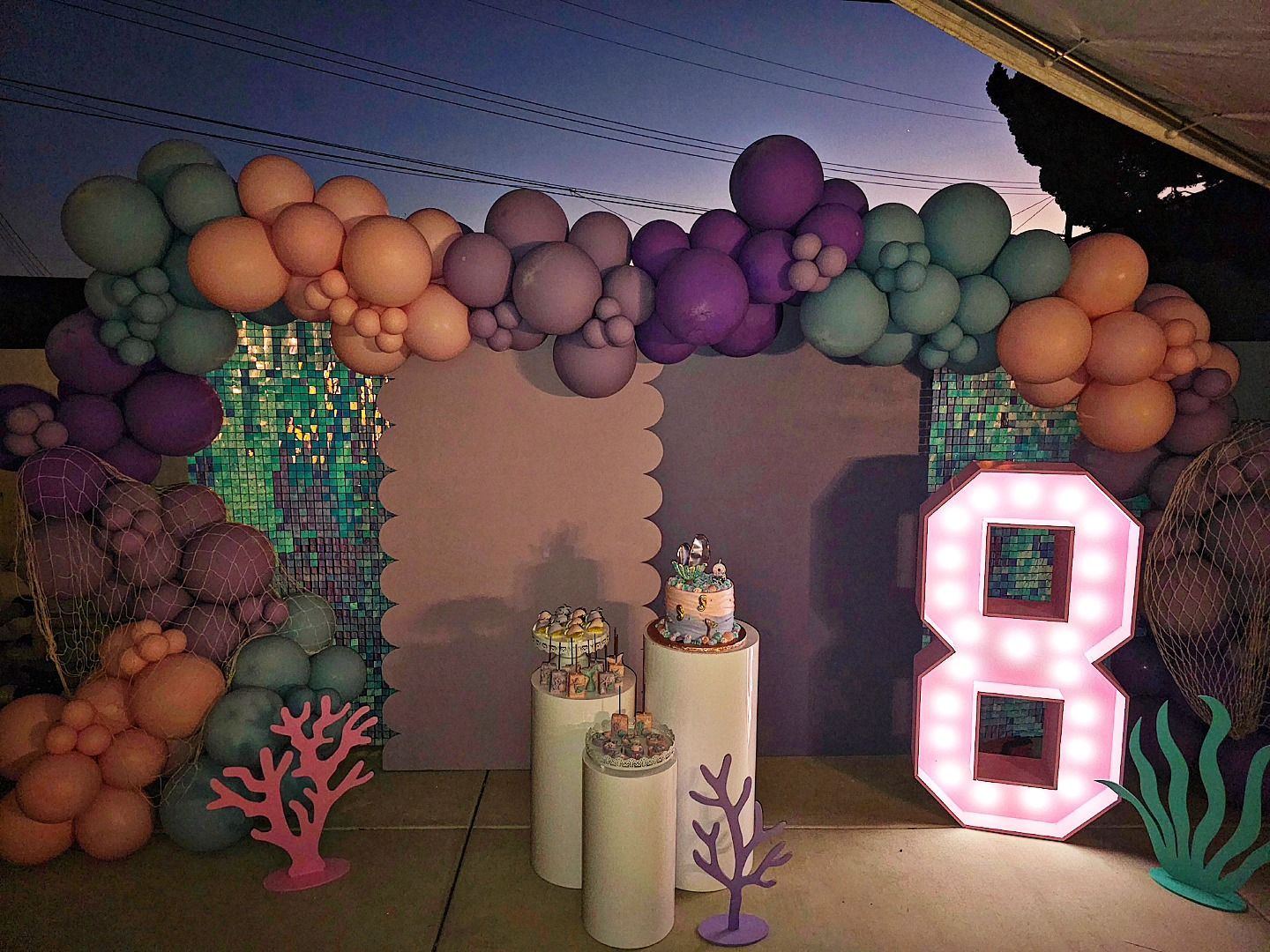Pastel balloon arch over a party dessert table with a glowing pink marquee number 8 and sea-themed decorations