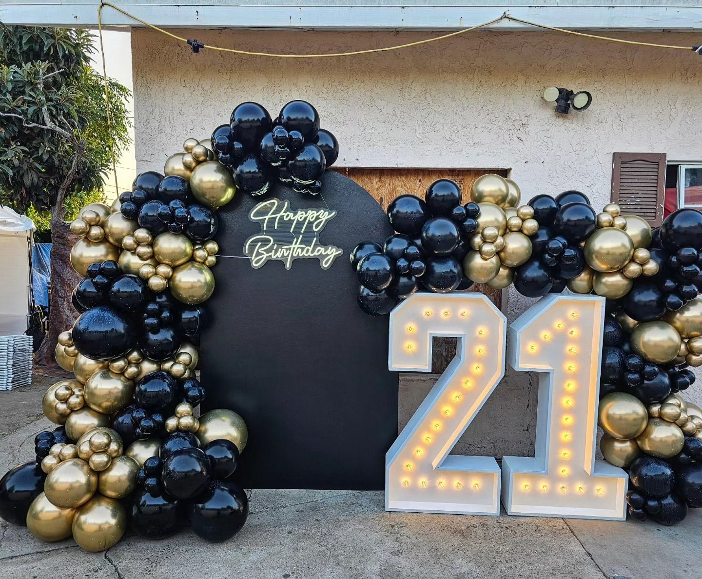 Black, gold, and white 21st birthday balloon backdrop with lit marquee numbers outdoors