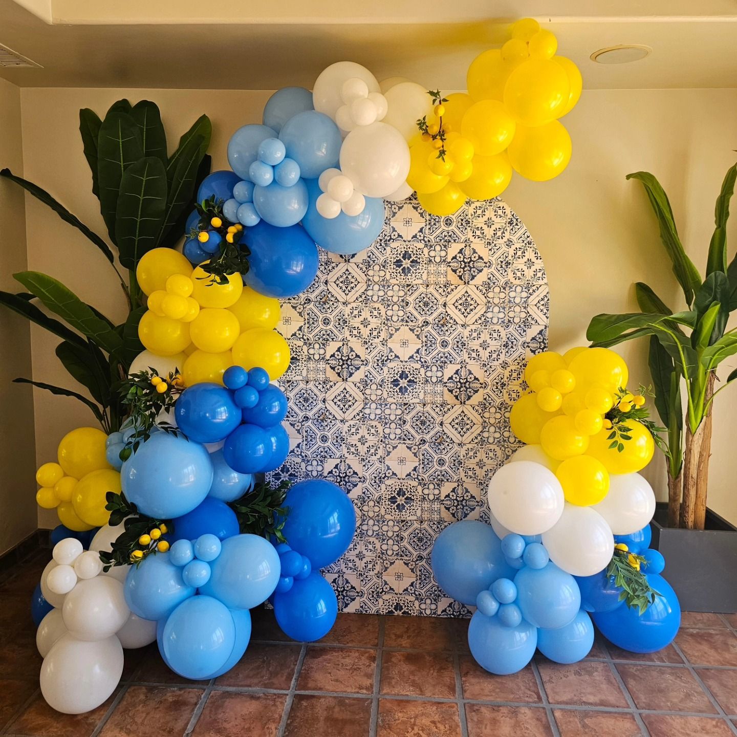 Blue, yellow, and white balloon clusters framing a tiled backdrop with potted plants on both sides