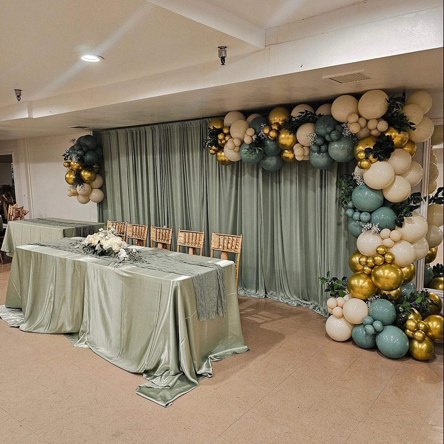 Decorated banquet table beside sage curtains and a gold, cream, and teal balloon arch in a hall
