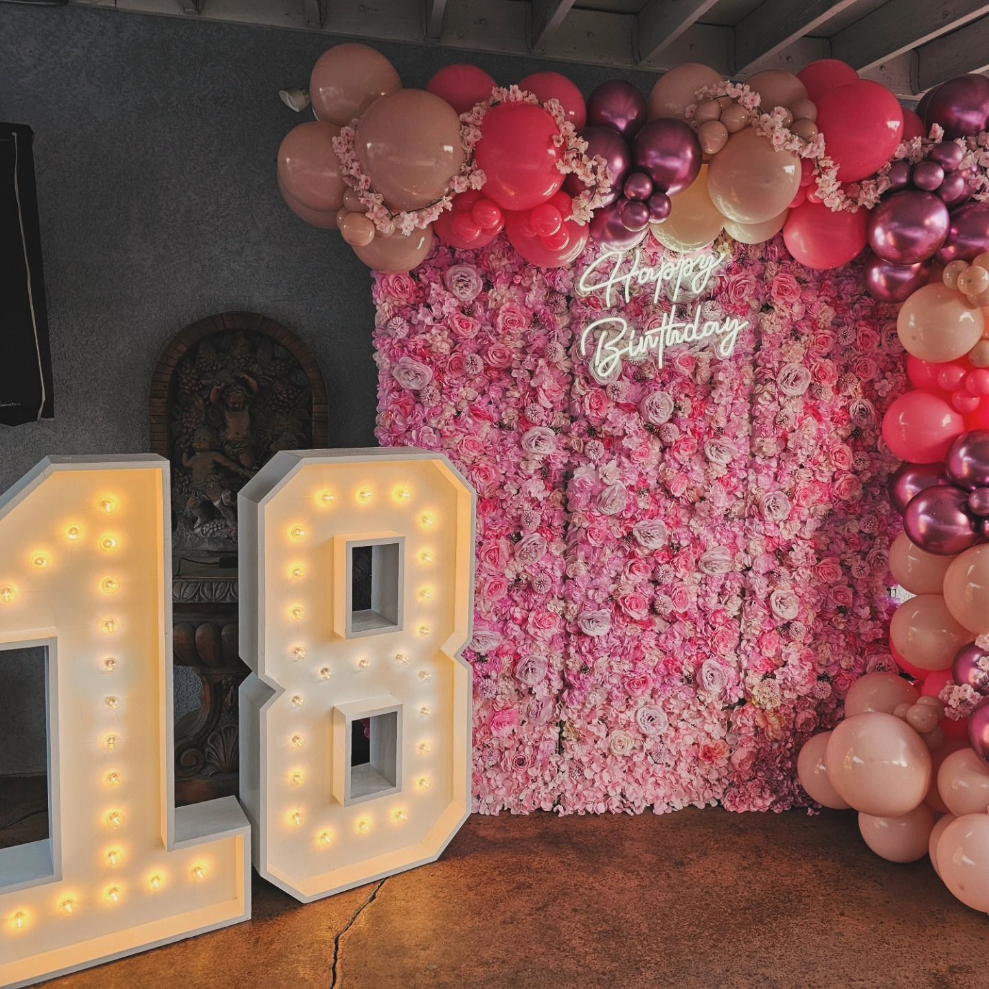 18th birthday party backdrop with pink floral wall, balloon arch, and lit marquee numbers 18