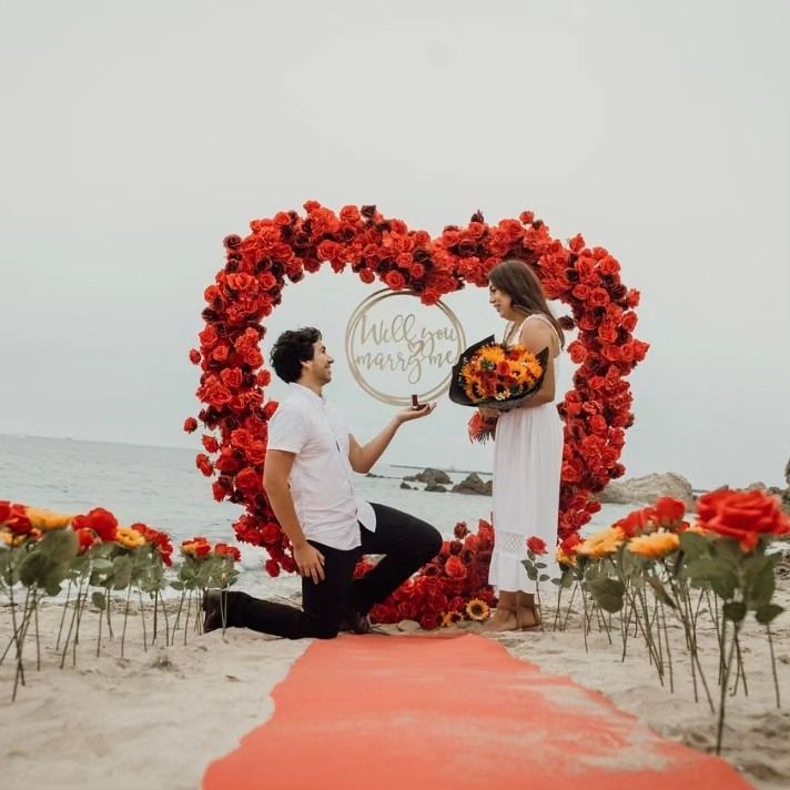 Beach proposal with red floral arch, orange aisle, and couple exchanging flowers by the sea