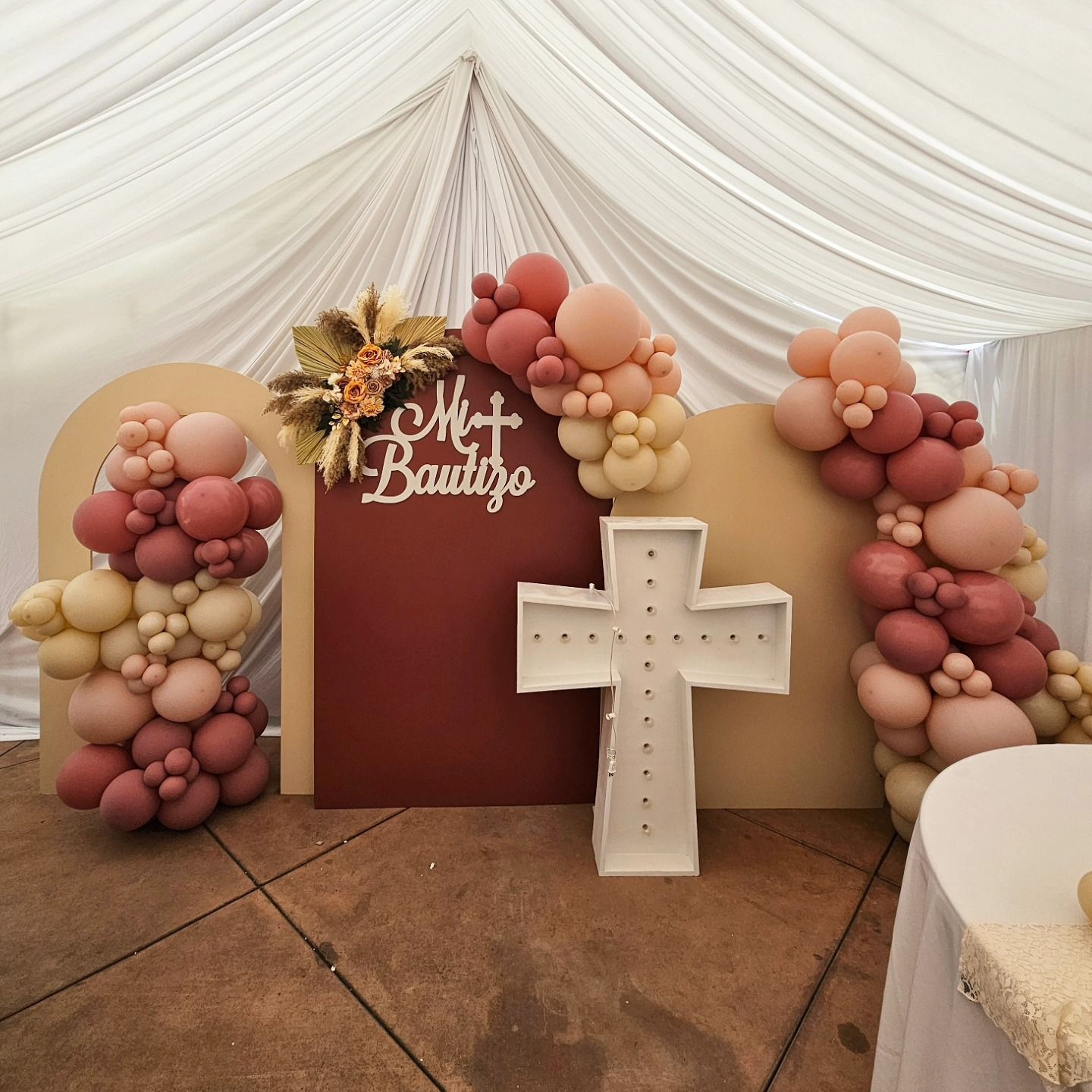 Decorated event backdrop with pink balloon arches, red and beige panels, flowers, and a white cross display
