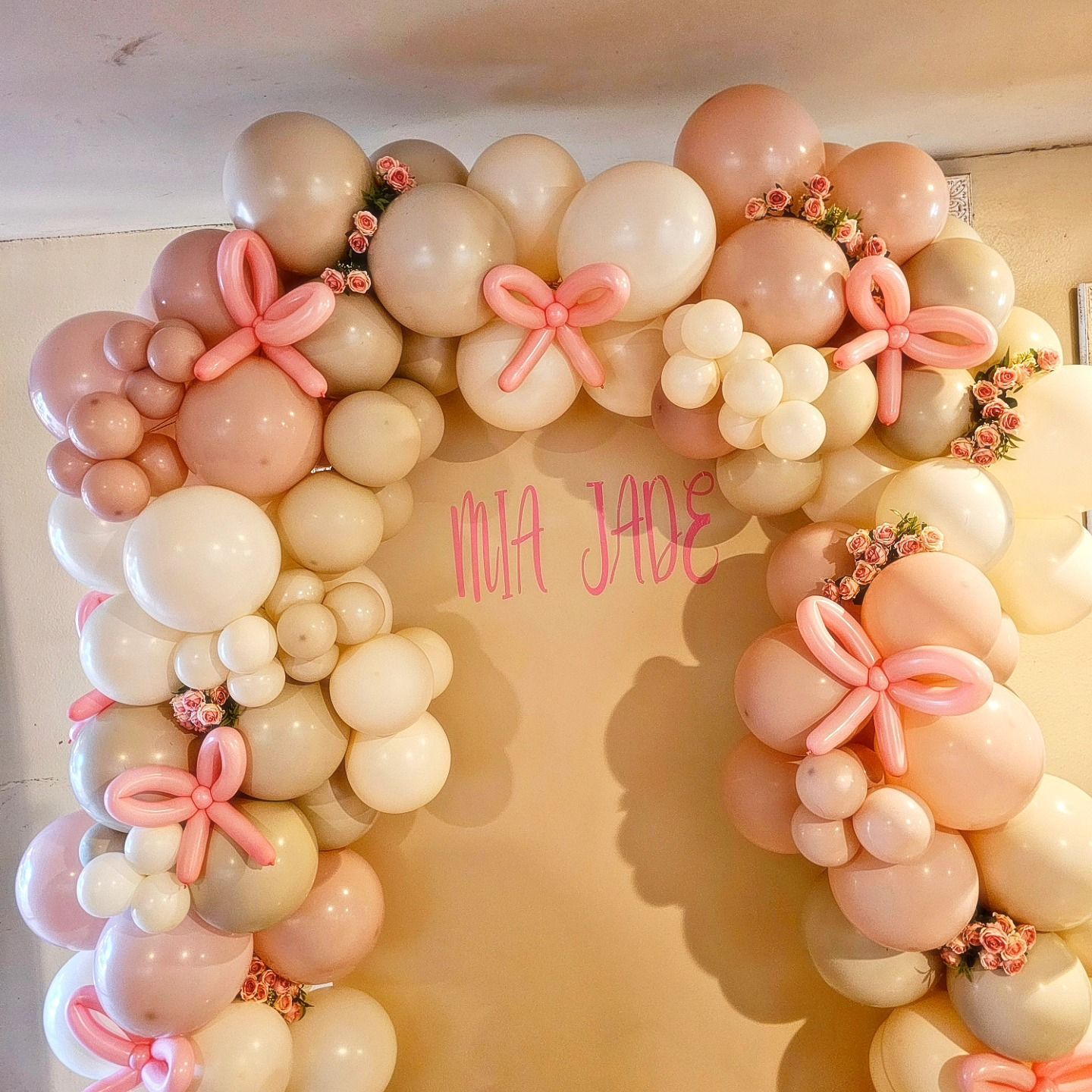 Pastel balloon arch with pink flower accents and center name text on a beige background