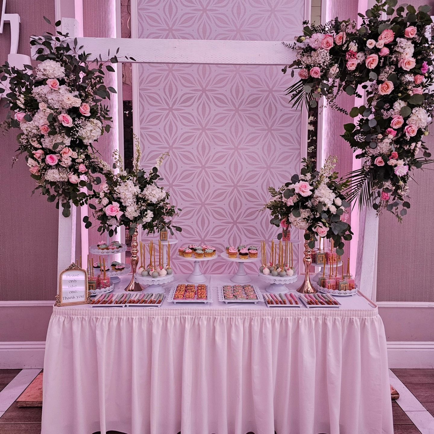 Pink floral dessert table with white draping and glassware against a pink patterned backdrop