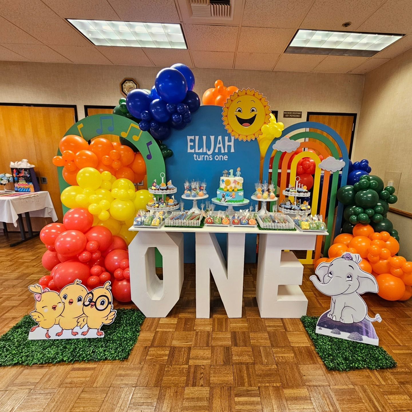 Colorful first-birthday party backdrop with blue “ELIJAH” sign and large white “ONE” letters with balloons and cartoon
