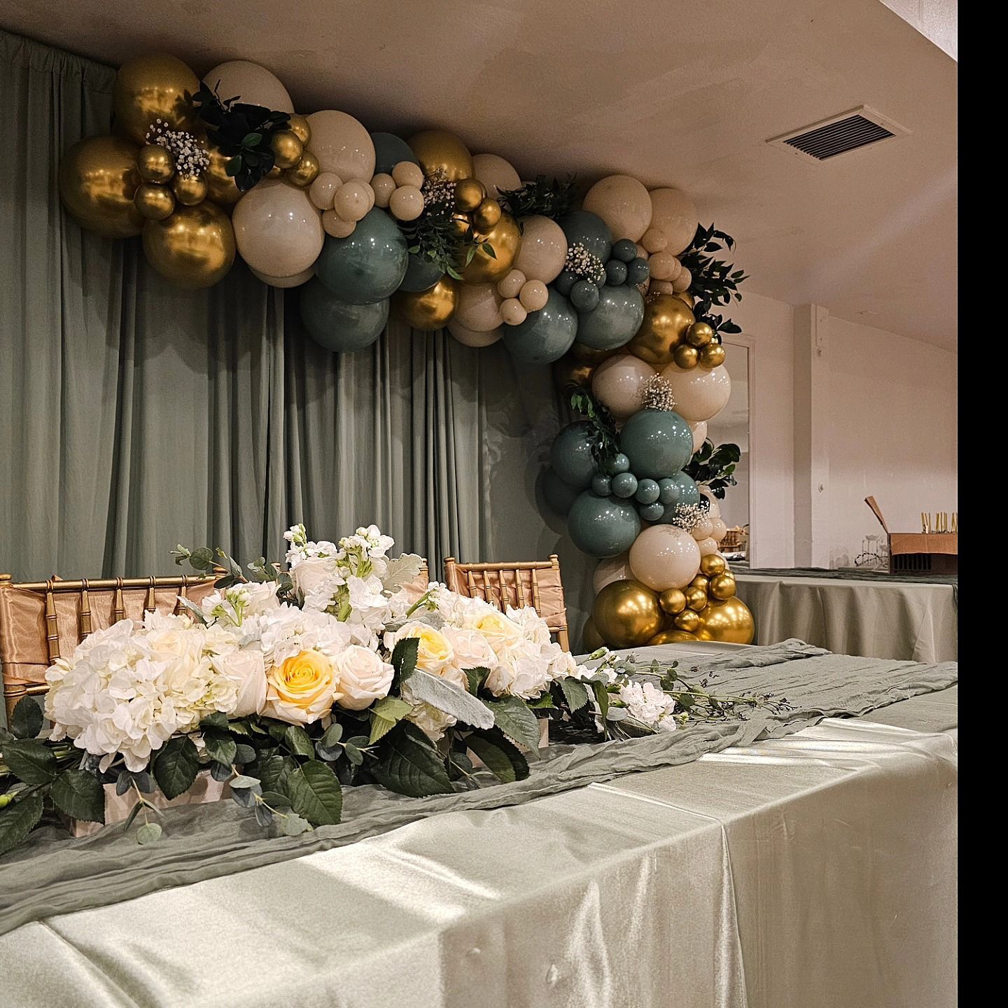 Elegant event table with white flowers and a gold, cream, and sage balloon arch backdrop