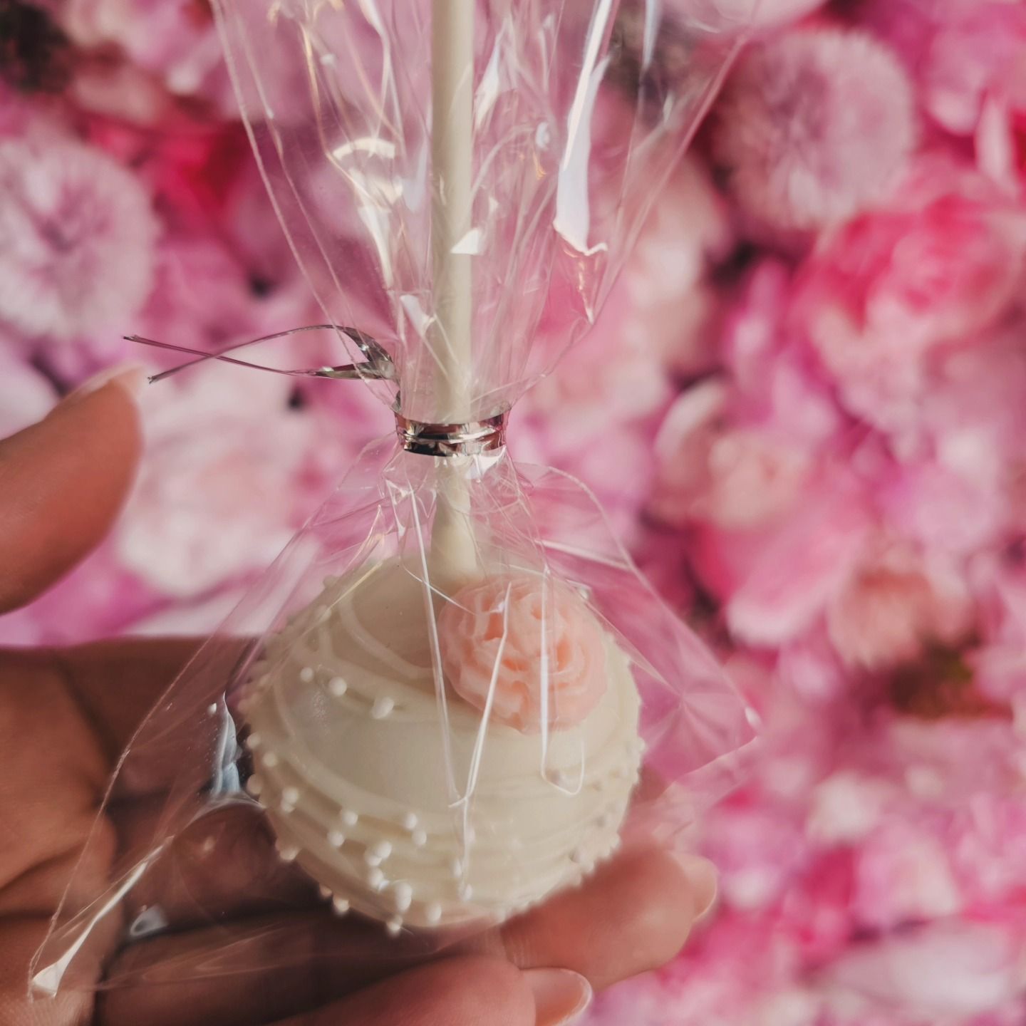 Hand holding a wrapped white cake pop with pink rose topper against a pink floral background