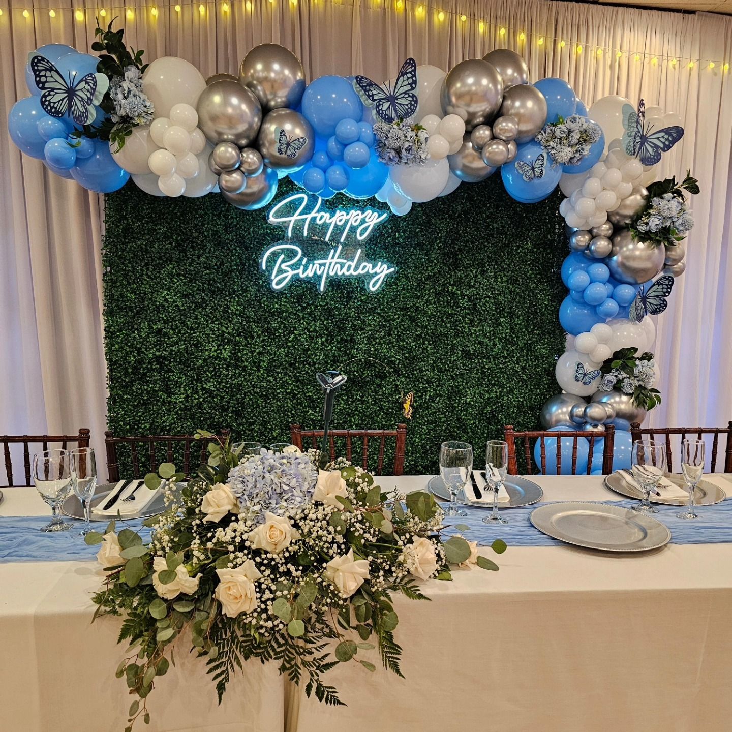 Birthday party table with white florals, blue and silver balloons, and a glowing “Happy Birthday” sign on a greenery wall