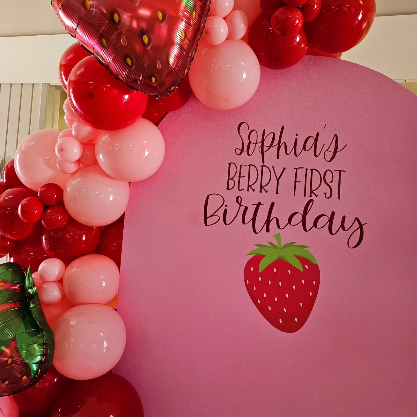 Pink strawberry birthday backdrop with red and pink balloon arch and “Sophia’s Berry First Birthday” text