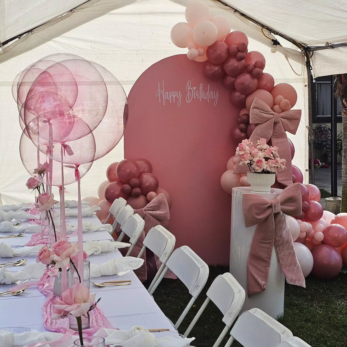 Pink balloon party backdrop with round sign, floral arrangement, and long decorated table under a tent