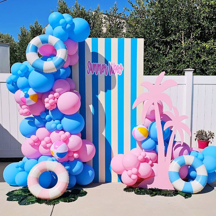 Blue, pink, and white balloon birthday display with striped backdrop and palm tree decor outdoors