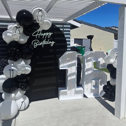 Black-and-white birthday balloon display with large silver “16” numbers outdoors under a pergola