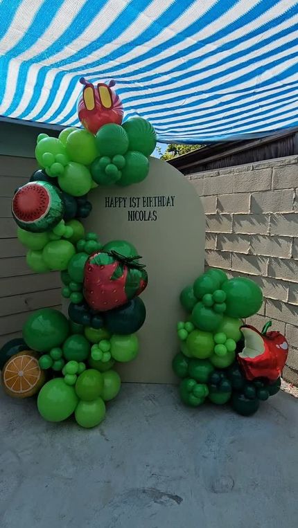 Green balloon arch around a stone sign under a blue-and-white canopy outdoors