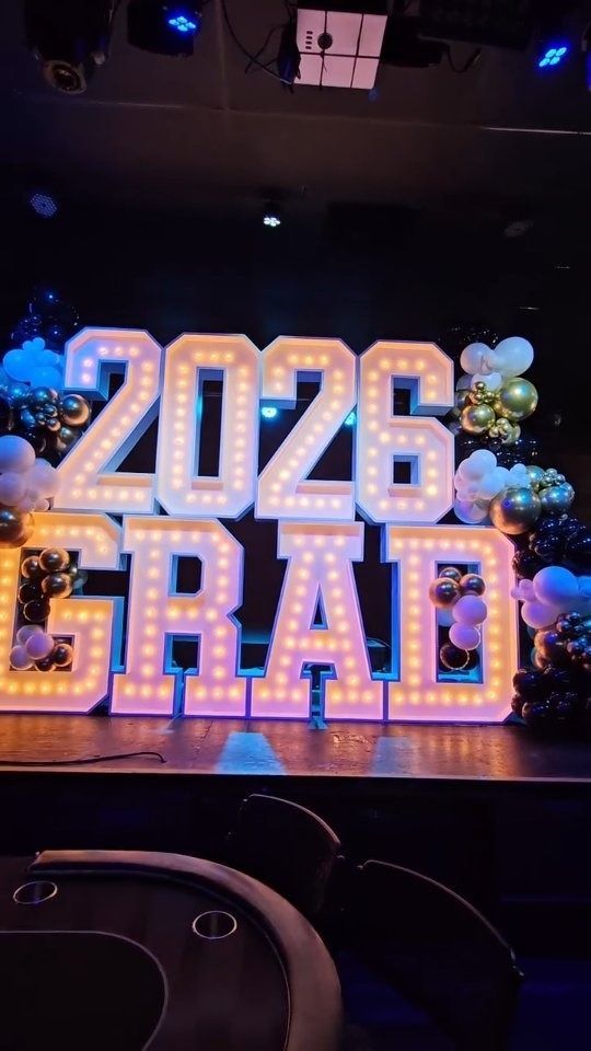 Lit “2026 GRAD” marquee letters on a stage with colorful balloon decorations.