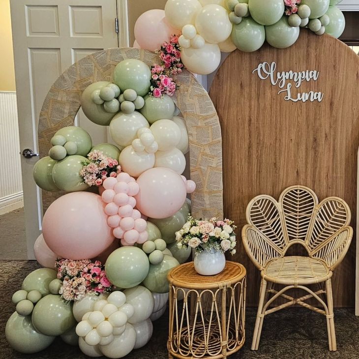 Pastel balloon arch and wicker chair display for a baby shower, with a small table and floral accents