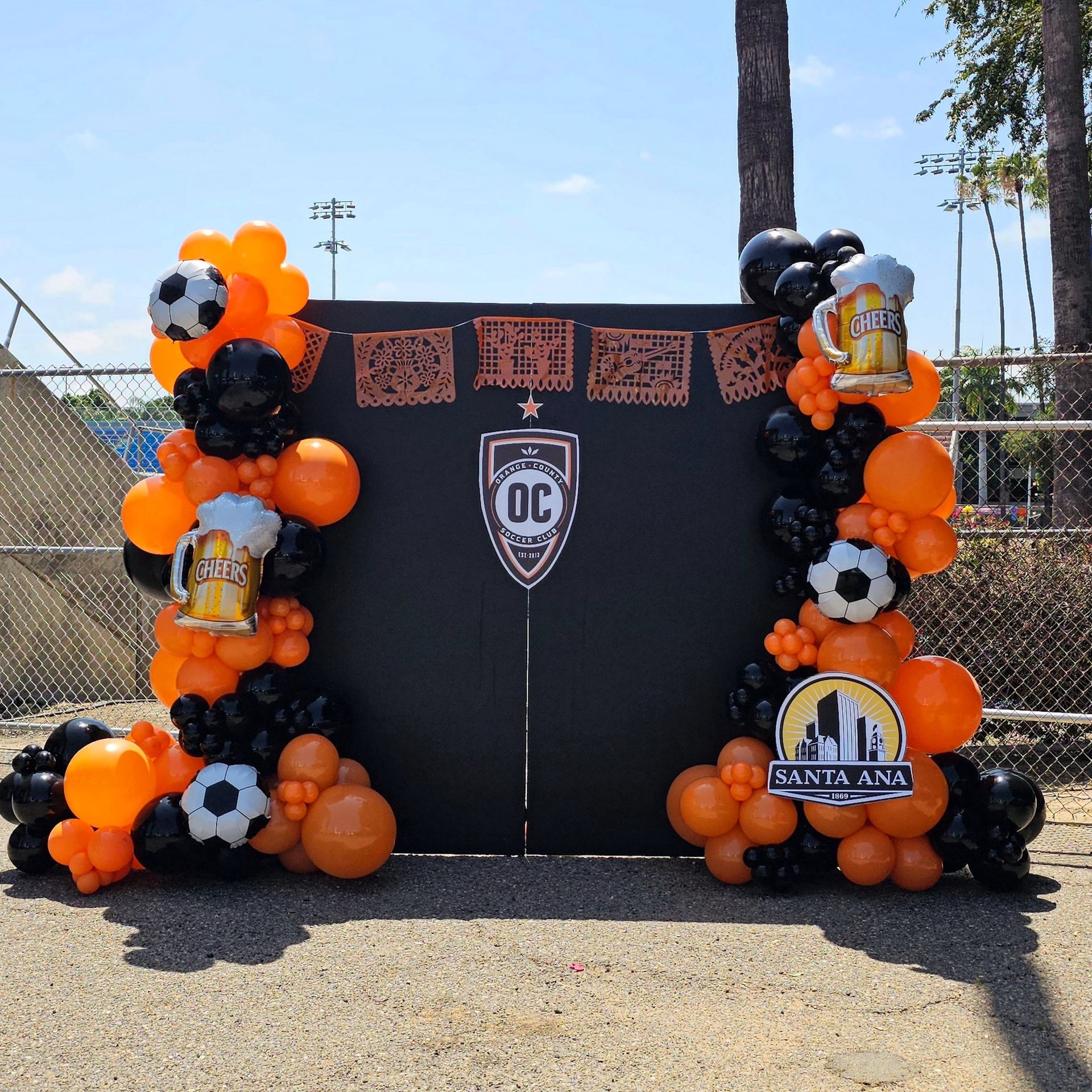 Orange-and-black balloon arch framing a sports backdrop with soccer balls and Santa Ana signage outdoors