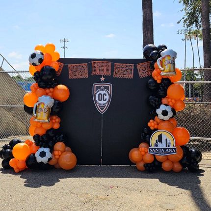 Orange-and-black balloon arch framing a sports backdrop with soccer balls and Santa Ana signage outdoors