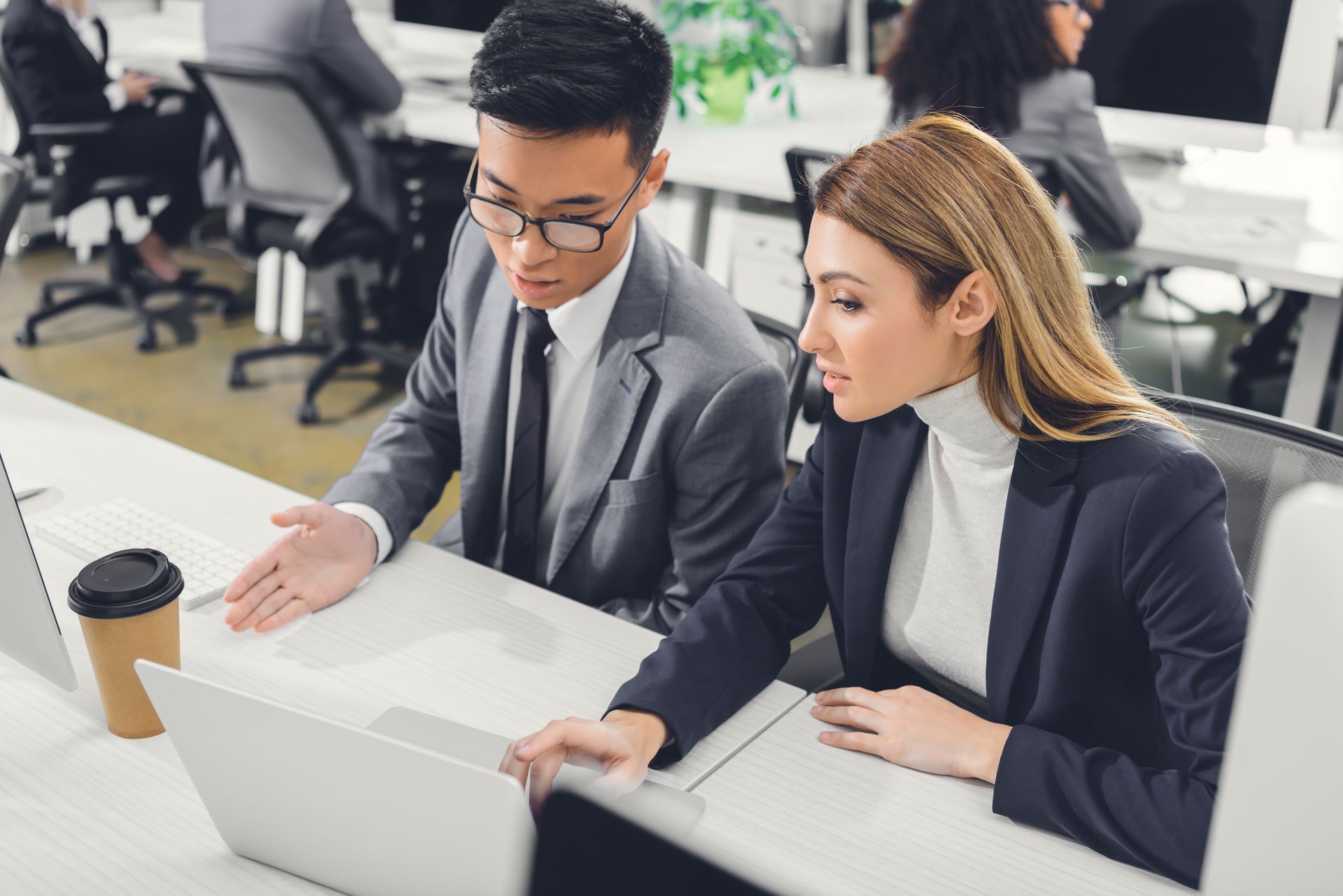 Two people in business attire look at a laptop in an office setting.