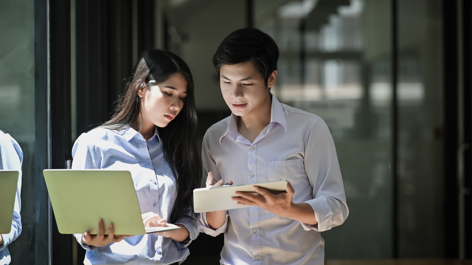 Two young people looking at a laptop and tablet, possibly collaborating in an office setting.