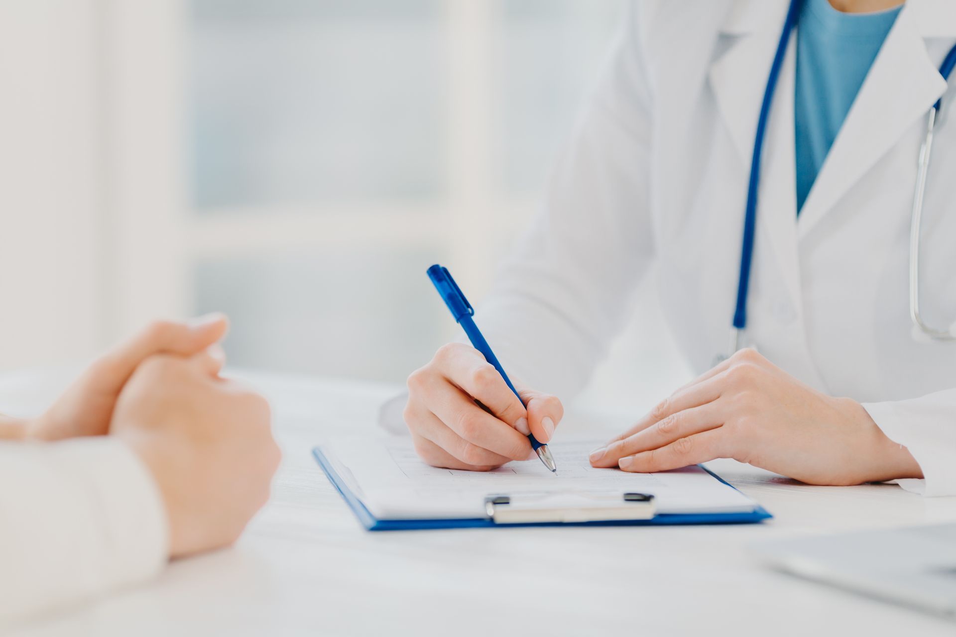 Doctor writing on clipboard, patient's hand visible. Doctor wears white coat and stethoscope.