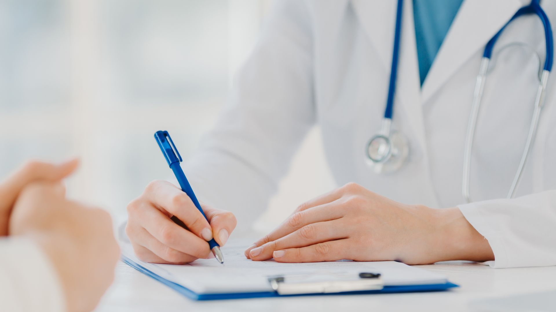 Doctor writing on clipboard, stethoscope around neck, patient's arm visible.