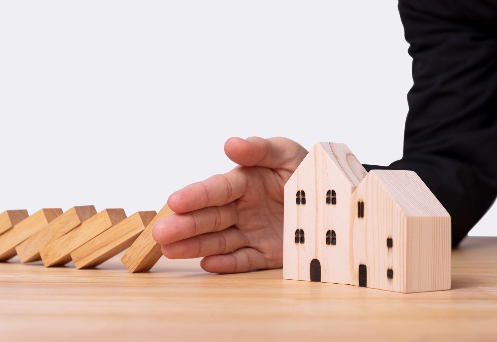 Hand protecting miniature houses from falling dominoes on a wooden surface.