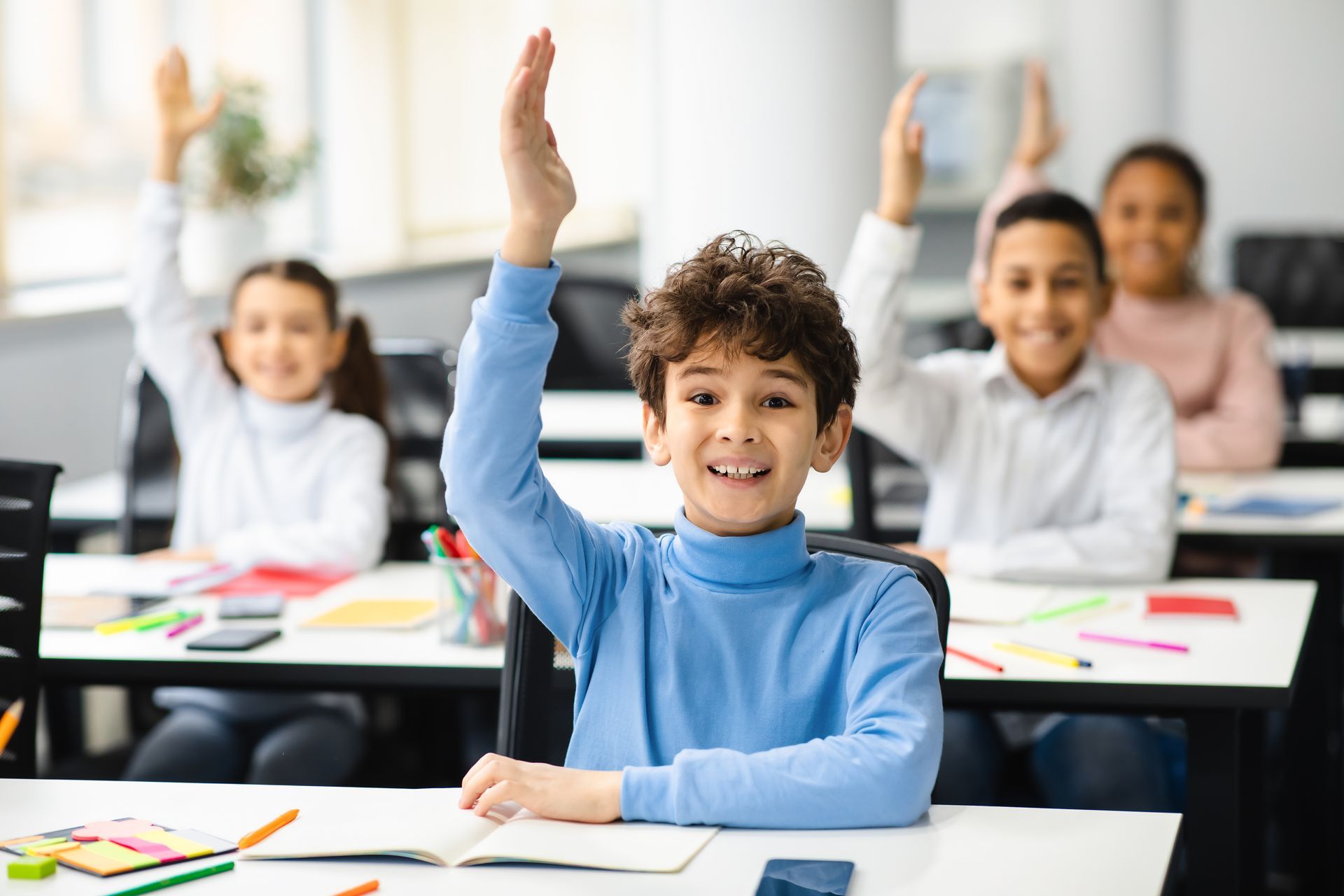 Children in classroom raise their hands enthusiastically, one boy smiling brightly.