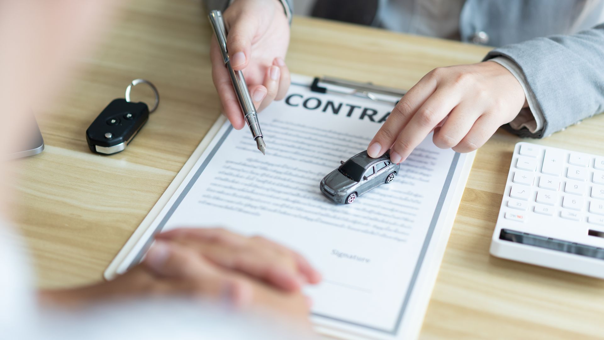 Person pointing to a car model on a contract, car key and calculator on desk.
