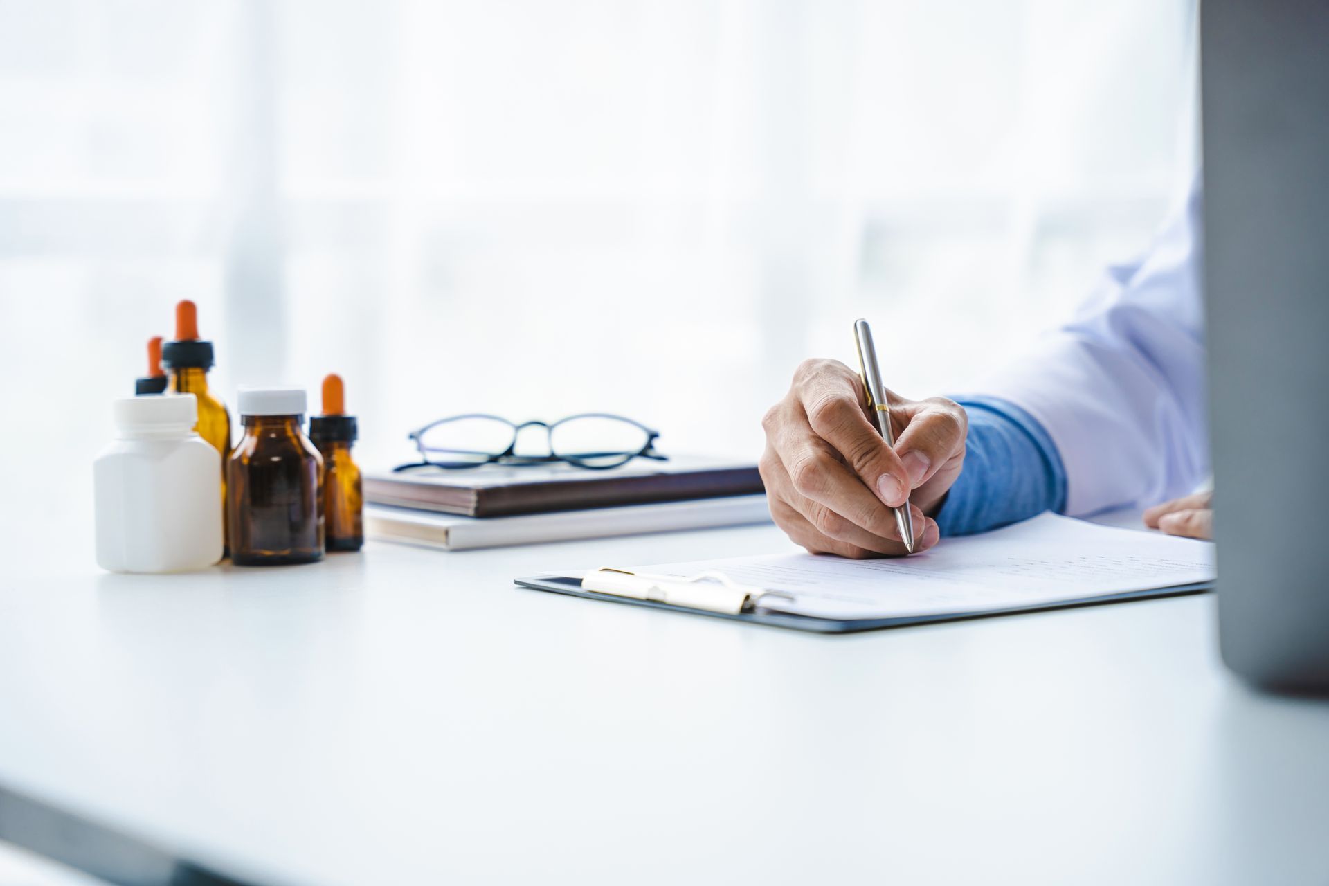 Doctor writing on clipboard at desk with medicine bottles, glasses, and a laptop.