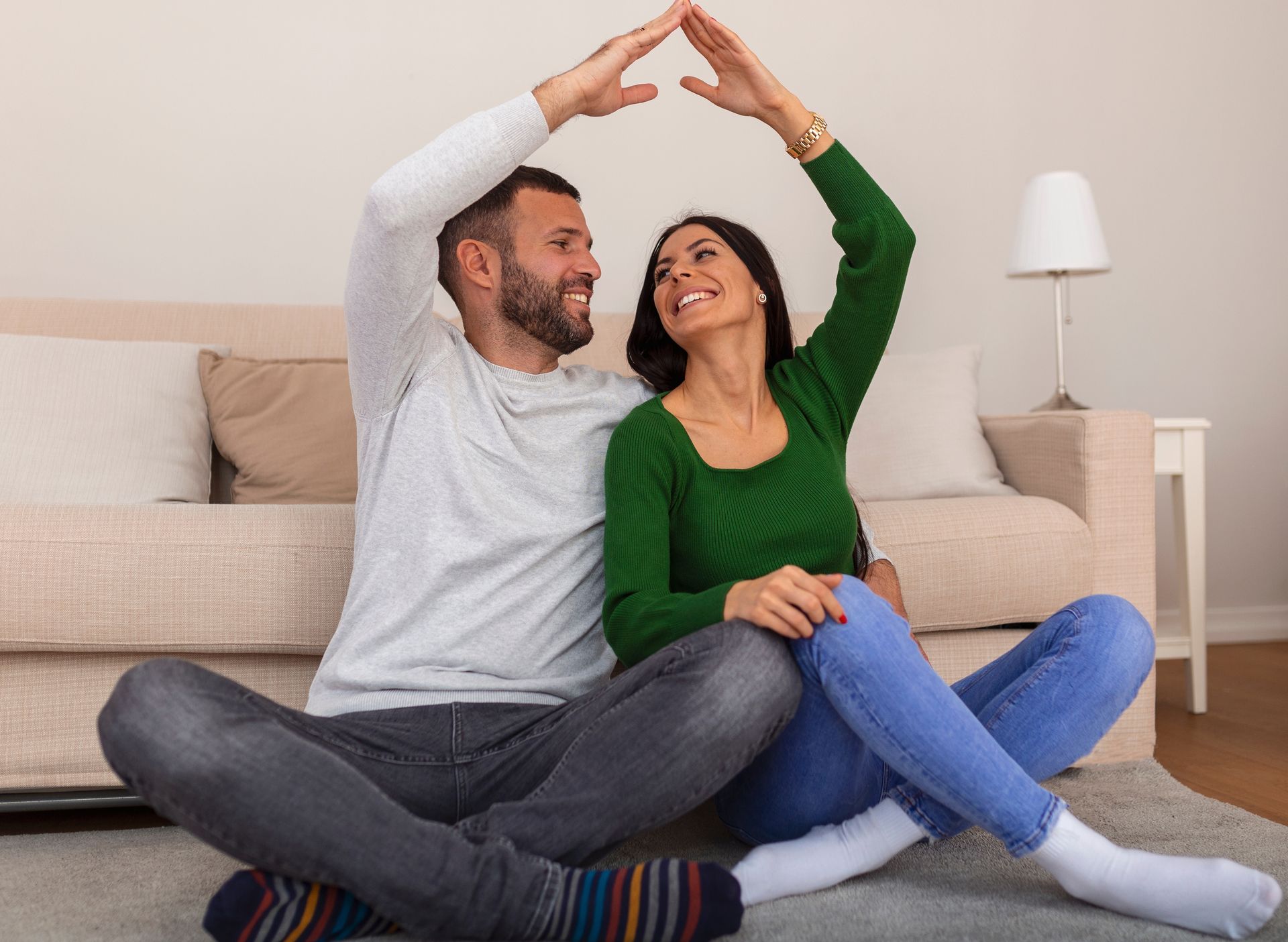 Couple sitting on floor, forming a house shape with their arms, smiling at each other.