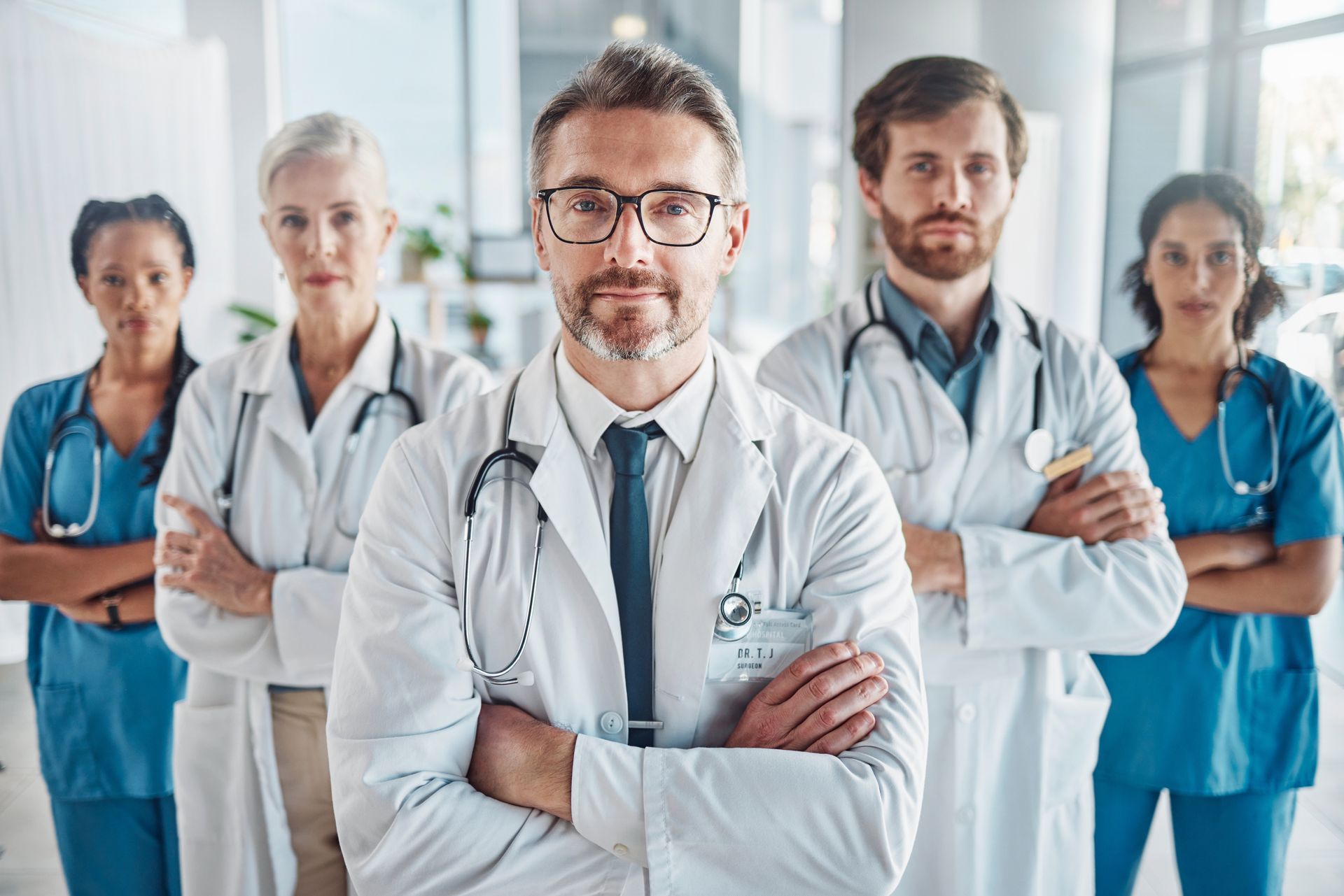 Medical team in lab coats with stethoscopes, arms crossed, looking at the camera. Bright setting.